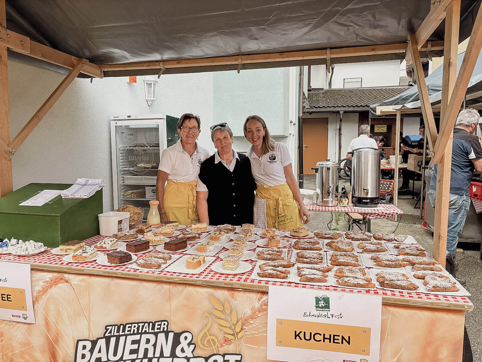 Drei Frauen stehen hinter einem Kuchenstand auf einem Bauernfest, mit verschiedenen Kuchen und Gebäck auf dem Tisch.