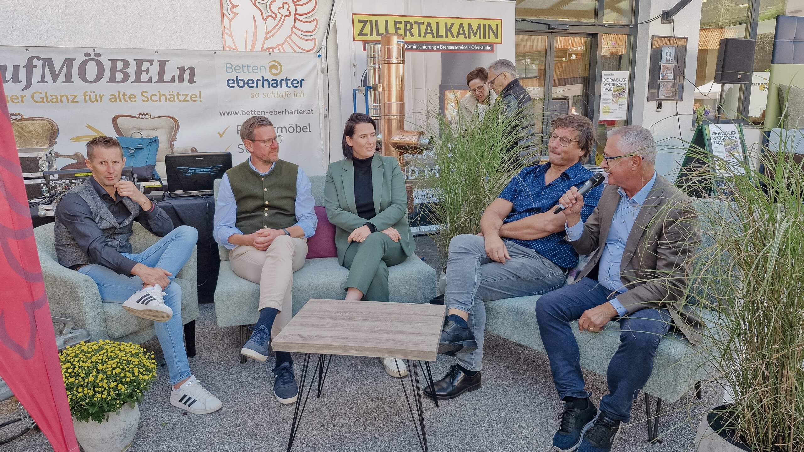 Fünf Personen sitzen auf Sofas bei einer Veranstaltung im Freien, umgeben von Werbeschildern und Pflanzen.